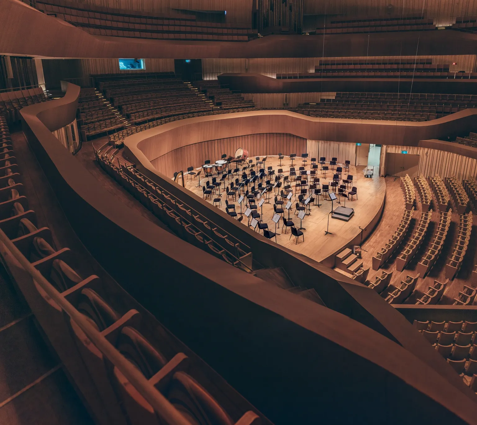 The stage of a modern timber concert hall, seen from the upper balcony. The chairs and music stands are set and no one is on the stage.